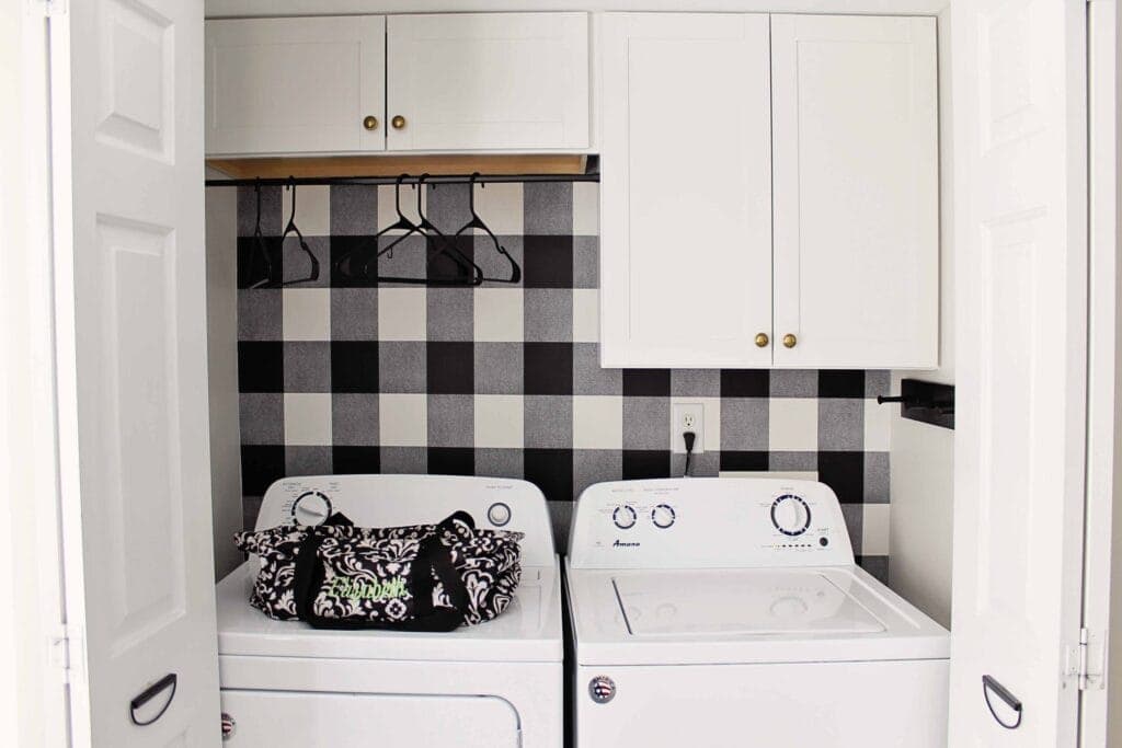 White washer and dryer in a laundry closet with a black-and-white checkered wall, overhead cabinets, and a hanging rod.